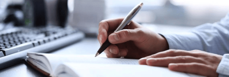 A man writes in a planner with a pen while sitting at a desk. 