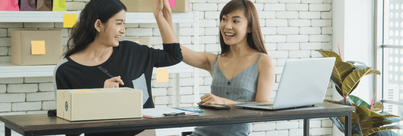 Two woman high-fiving while sitting on a table with a computer and a box