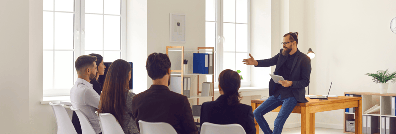 A man giving a presentation to a group of 5 people sitting in front of them and looking at a small screen