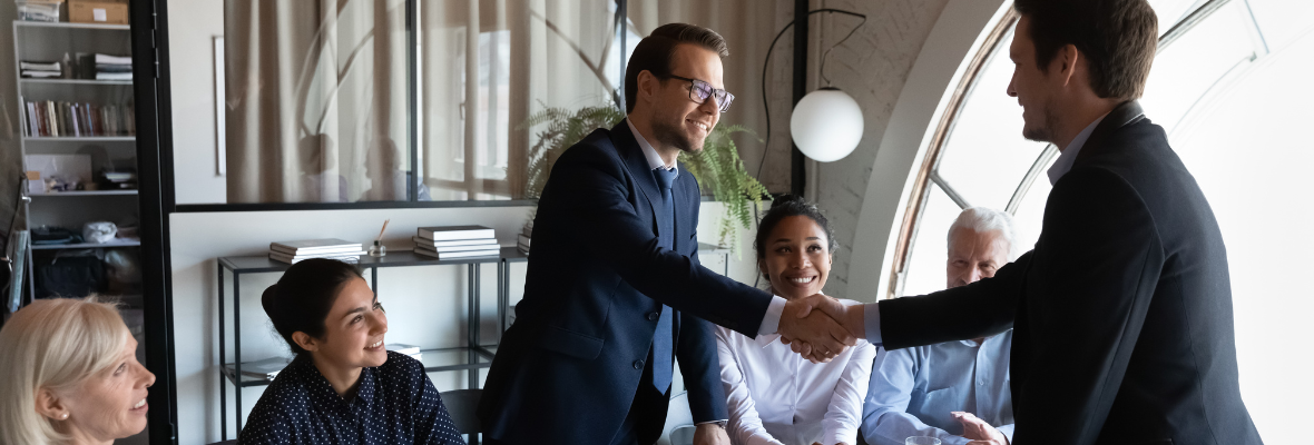 A sales team gathered in an office table while two man are shaking their hands as in closing a deal