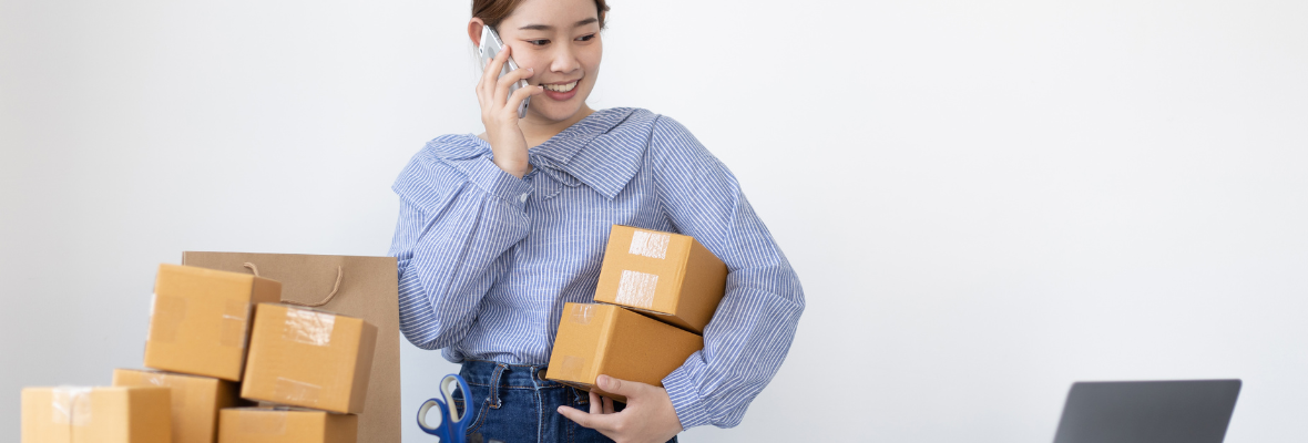 Asian woman holding multiple boxes while talking on the phone and looking at her laptop