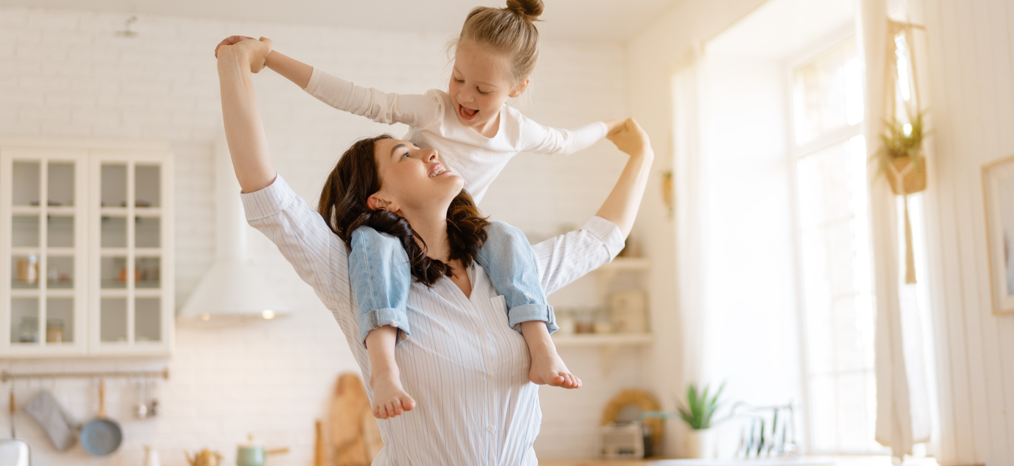 Mother joyfully carrying daughter on shoulders in a bright kitchen, both smiling and holding hands up, capturing a happy and playful moment.